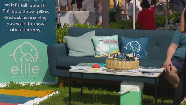 An outdoor event setting for Ellie Mental Health counseling with a blue couch and several throw pillows, a white table with various Ellie Mental Health branded items, and a large display sign asking people to pull up a pillow and ask questions about therapy.