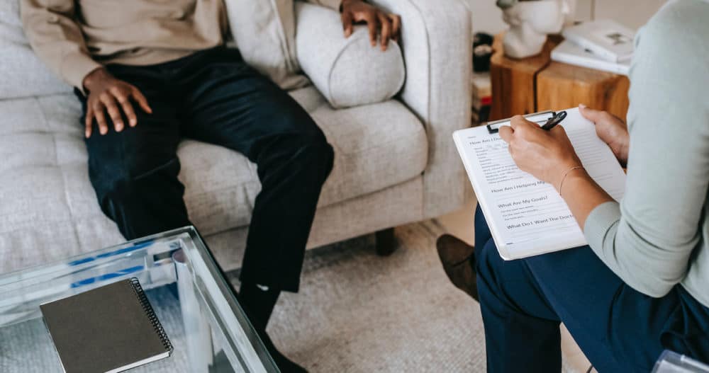 Black man sitting on a cream colored couch across from a woman therapist holding a clipboard taking notes