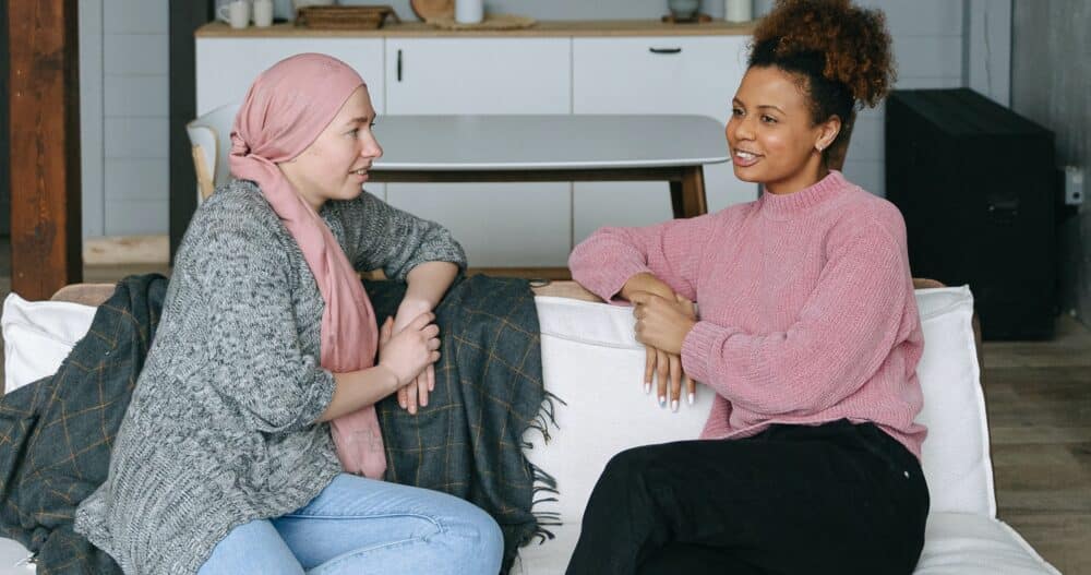 A woman sitting on a couch wearing jeans, a gray and black sweater, and a pink scarf on her head speaks with another woman sitting next to her who's dressed in dark pants and a pink sweater.