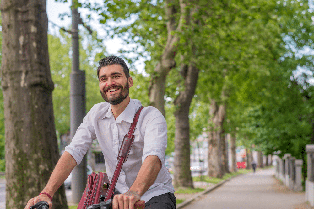 Man with a beard wearing a button up shirt and crossbody bag riding a bike and smiling in front of some trees in the city