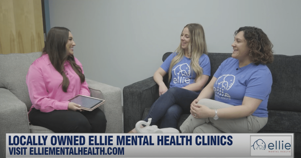 A woman with long dark hair sitting in a grey chair interviewing two women in Ellie Mental Health tshirts who own local Ellie Mental Health clinics.
