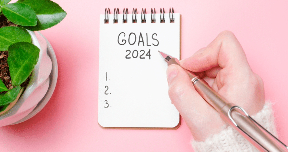 A hand holding a pen writing down goals for 2024 on a notepad on a pink desktop with a small green plant nearby.